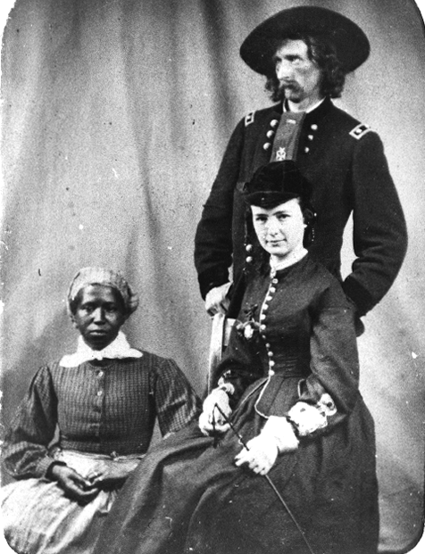 Historical photograph of General George Armstrong Custer standing behind his wife Elizabeth Bacon Custer and Eliza, seated together, circa 1865.